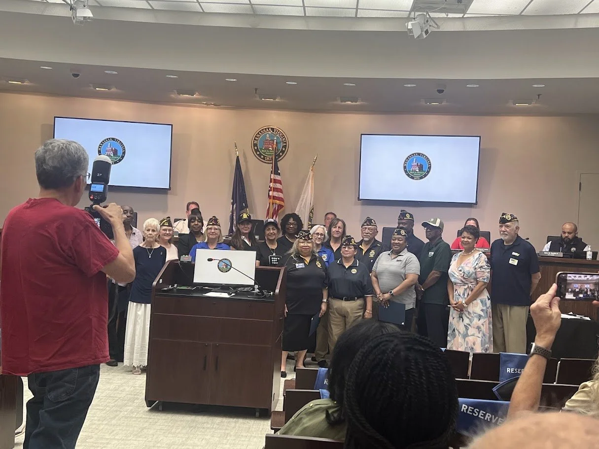 Group of people standing in a meeting room or council chamber, some wearing uniforms and hats, with flags and a seal on the wall behind them, as a person in a red shirt takes a photograph.