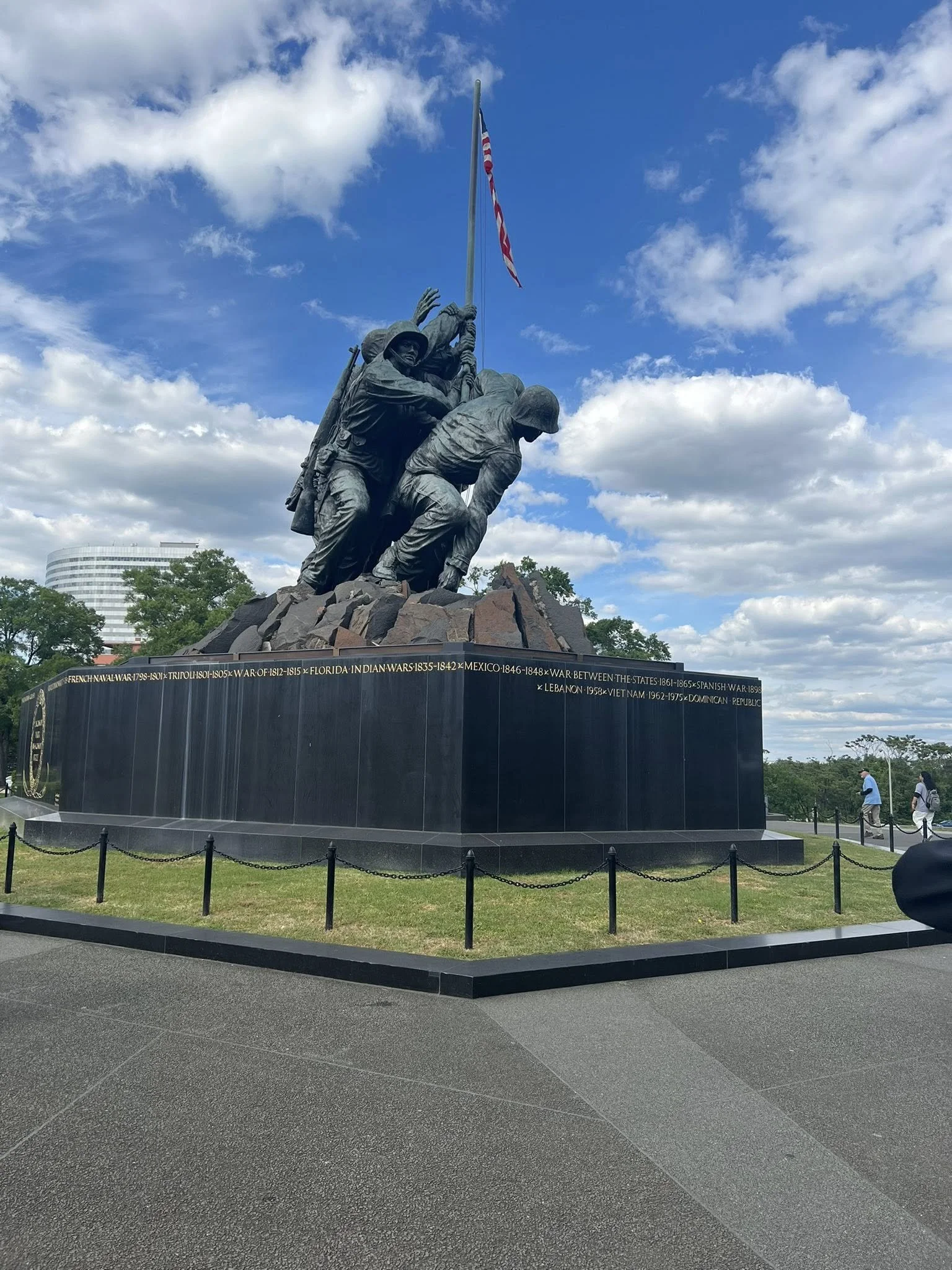 A monument featuring soldiers raising a flag, with a circular black base with inscriptions, outdoors on a partly cloudy day.