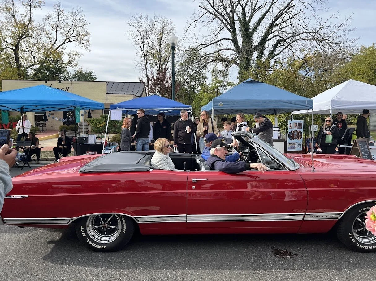 A red vintage convertible car with a black soft top, parked in front of a street fair with blue tents and people walking around.