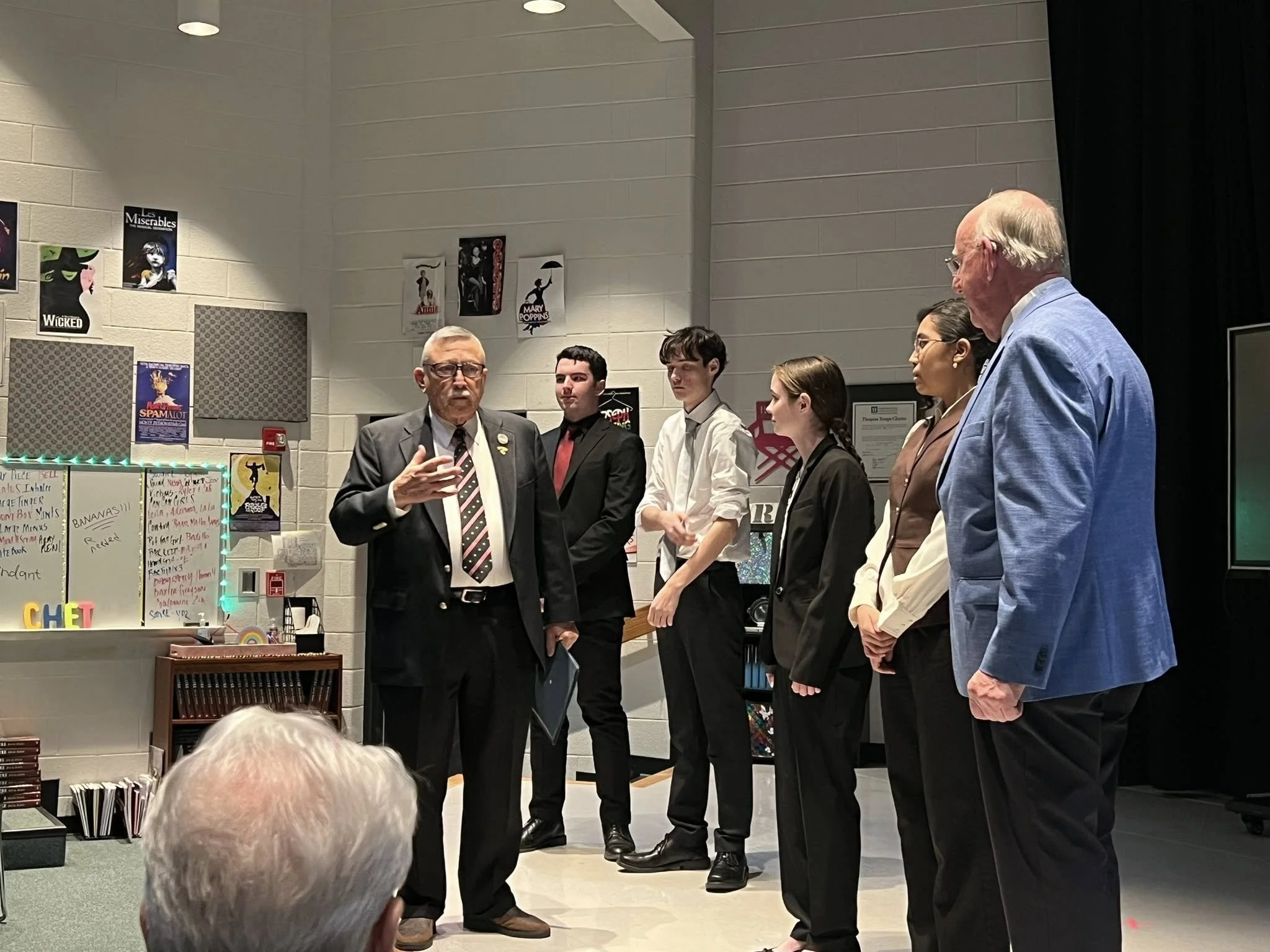 Group of six people standing in a classroom or presentation room, with posters on the wall and a board with writing, listening to a man in a suit who is speaking.