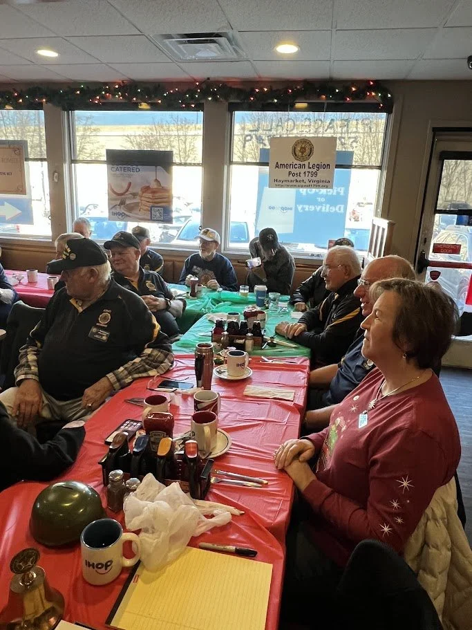 A group of people, mostly older adults, sitting at a long table covered with a red tablecloth in a restaurant or cafe decorated with Christmas garland. The people are wearing military or veteran pins, suggesting a veterans' gathering. There are cups,
