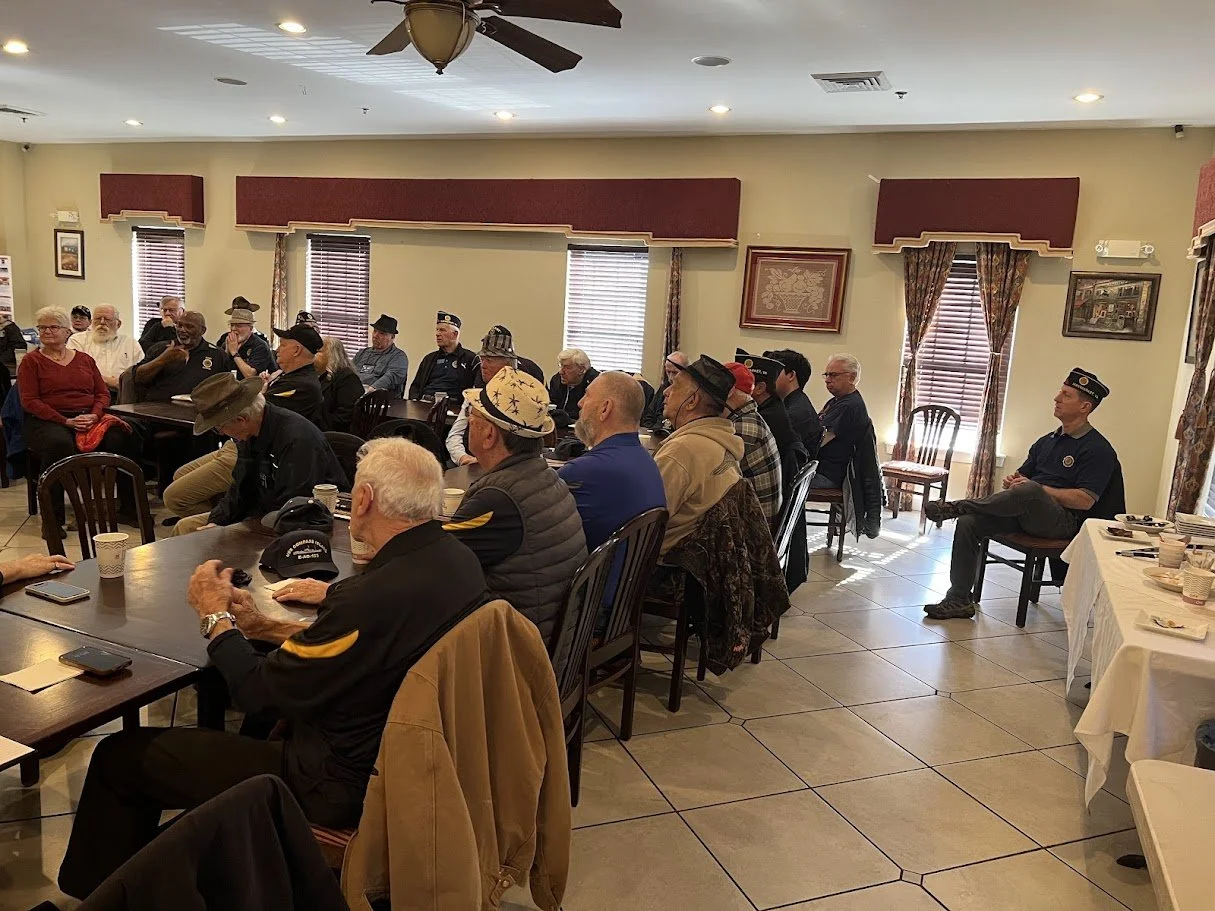 A group of elderly people sitting in a room, some wearing hats and uniforms, attending a meeting or gathering, with tables, chairs, and framed pictures on the walls.
