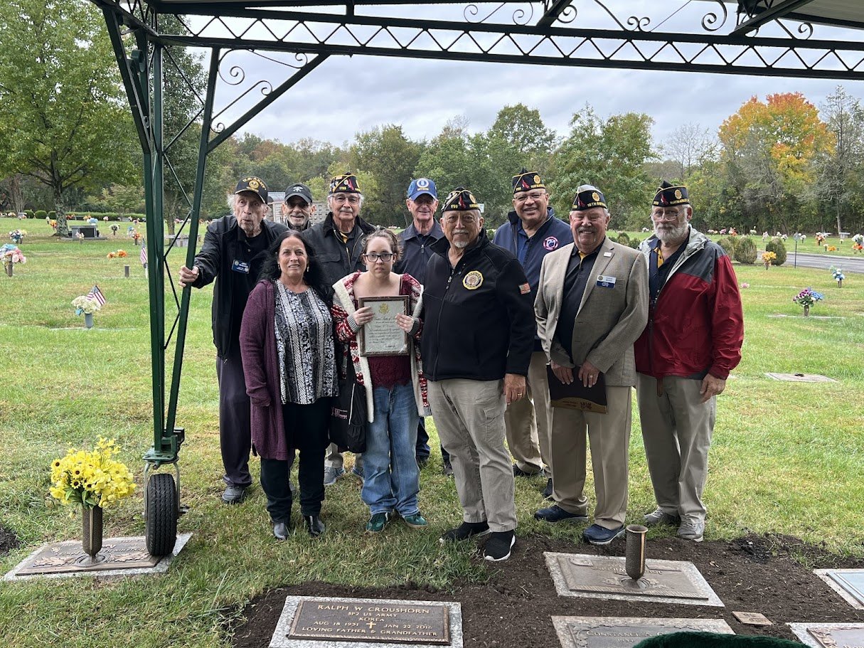 Group of people, including veterans with hats and a young girl holding a framed document, gathered under a memorial arch at a cemetery.