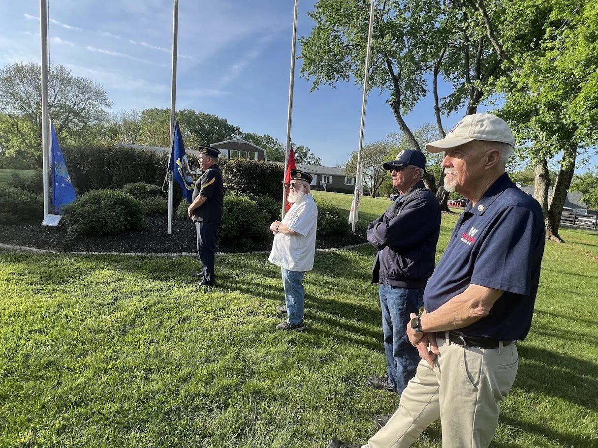 Four men standing outdoors in front of flagpoles, with flags including one with a blue field and another with red, participating in a memorial or ceremony. The setting is a grassy area with trees and a house in the background, under a partly cloudy s