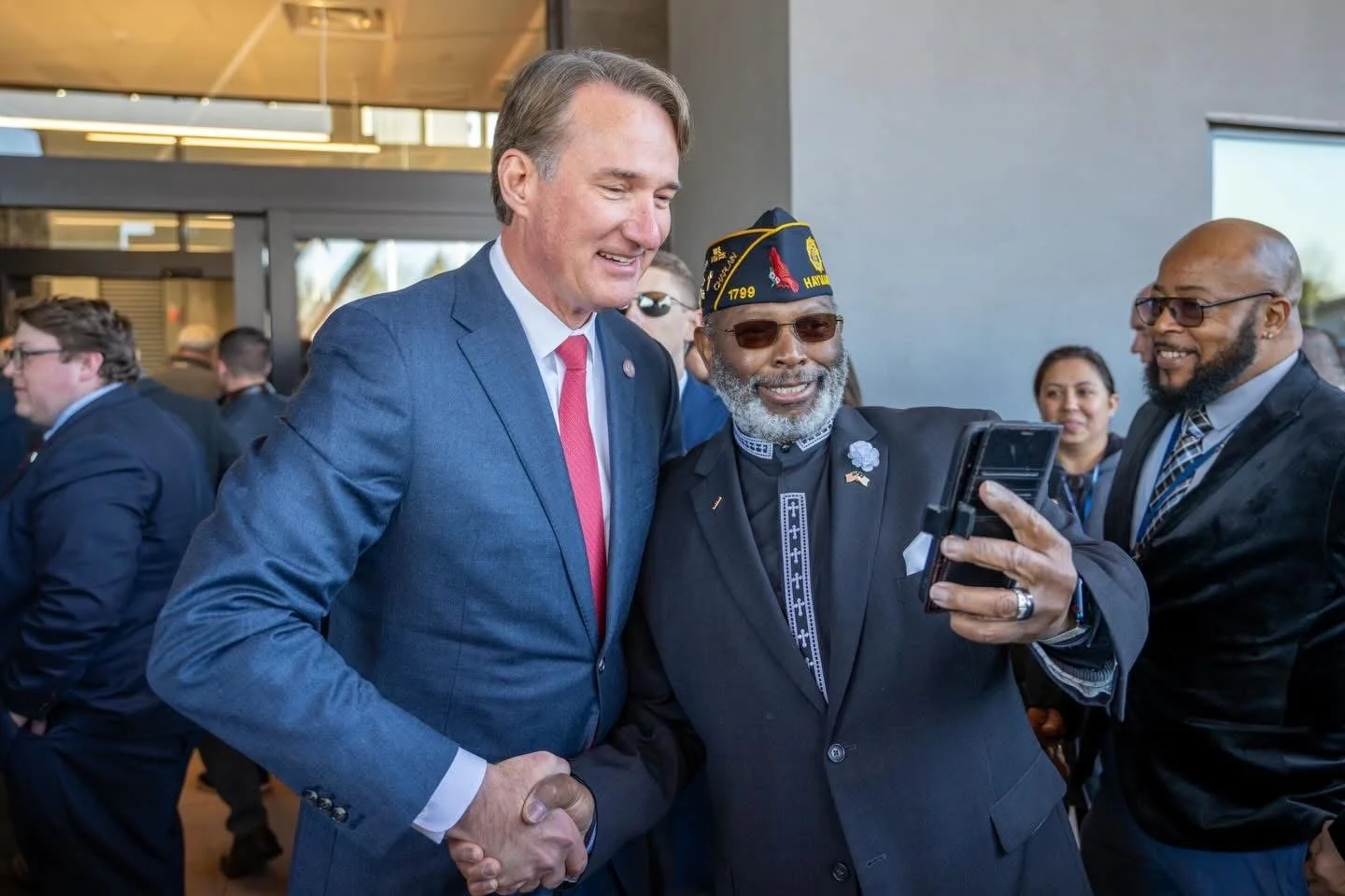 Two men, one in a gray suit and the other in a military cap and sunglasses, are smiling and looking at a smartphone together at an event, with several other people in the background.