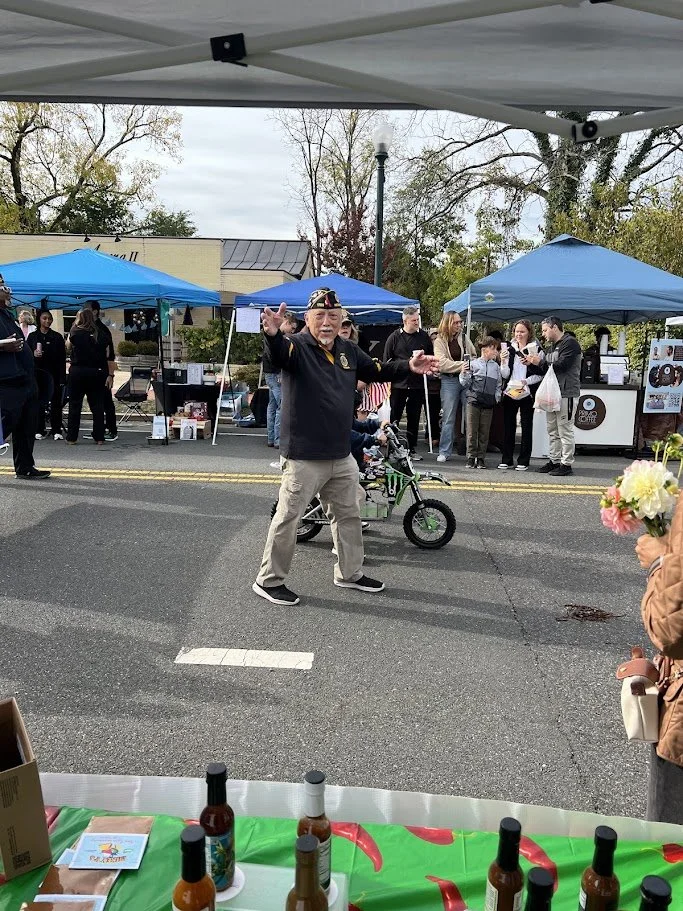 A man waving and smiling at a street market booth with tents and people behind him. There are bottles on the table in the foreground and a small child on a bicycle behind the man.