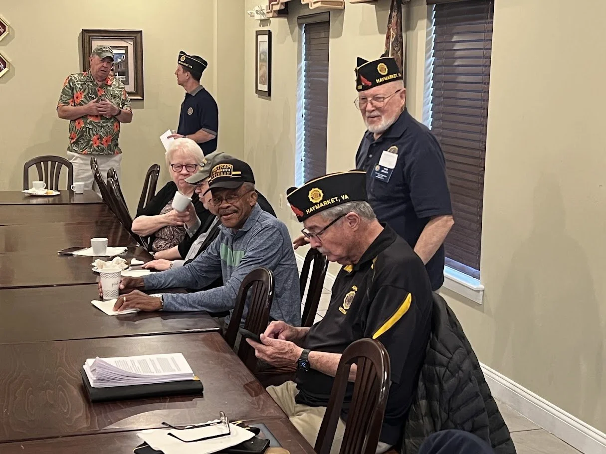 Group of veterans at a meeting, some seated, some standing, wearing military hats and casual clothing, in a room with framed pictures and windows with blinds.