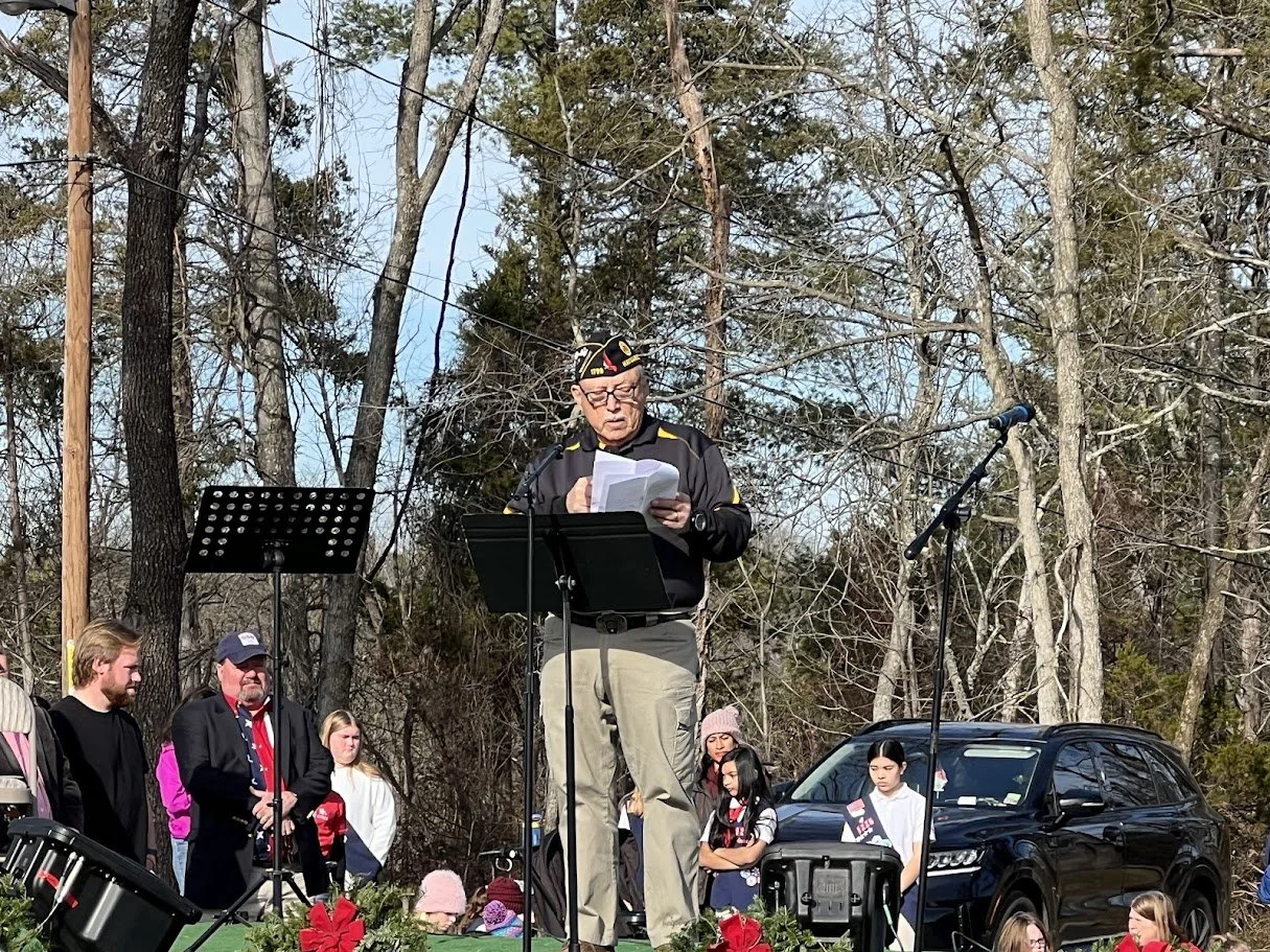 An elderly man wearing military attire and glasses speaking at an outdoor event, standing at a lectern with a microphone, surrounded by people and trees.