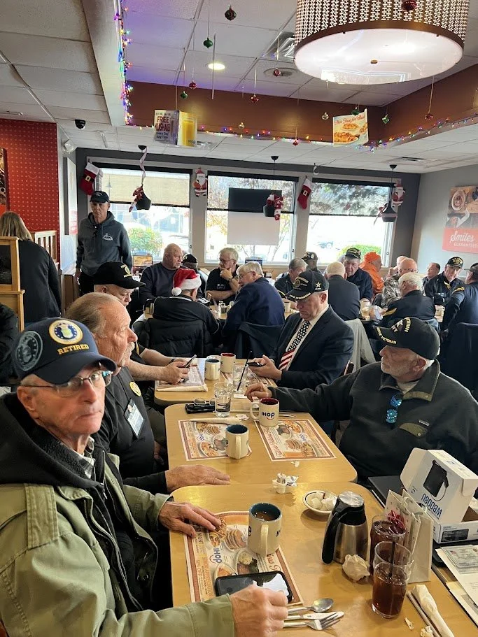 Veterans gathering at a restaurant during the holiday season, with some wearing hats and patriotic attire, decorated with Christmas ornaments and colorful lights.