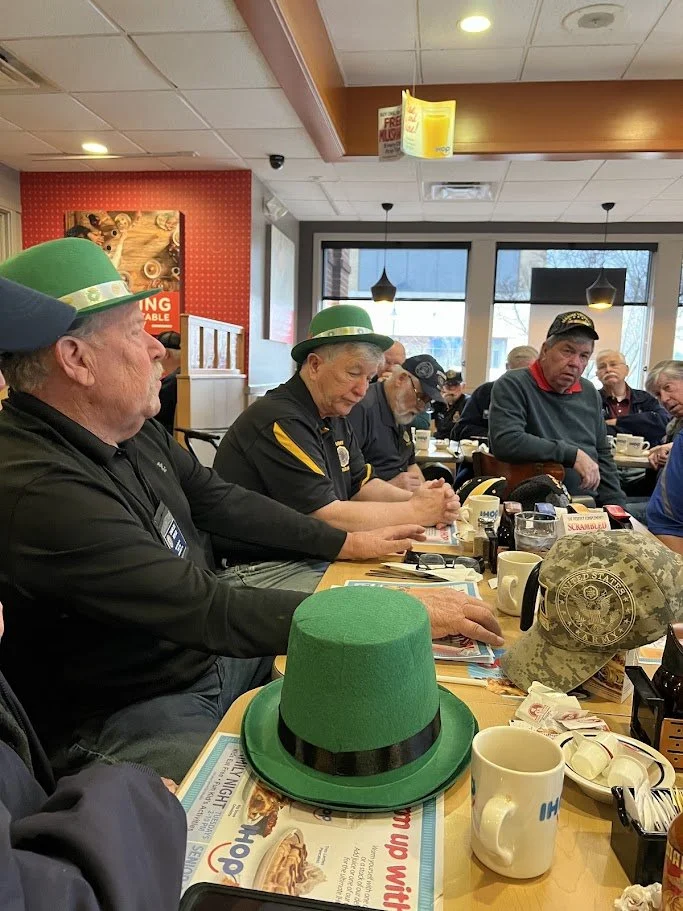 A group of men sitting at a restaurant table, wearing green hats, with several St. Patrick's Day themed items, including a green leprechaun hat and a camouflage hat with a military emblem, on the table.
