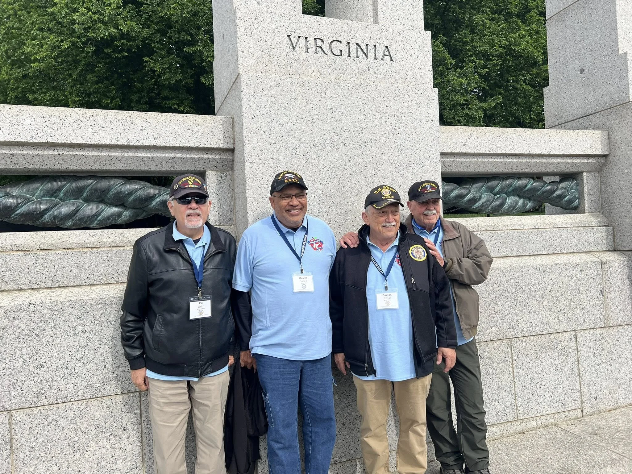 Four men standing in front of a stone monument with the word 'Virginia' engraved on it. The men are wearing jackets or shirts with military and patriotic patches, and they are smiling. The background includes green trees.