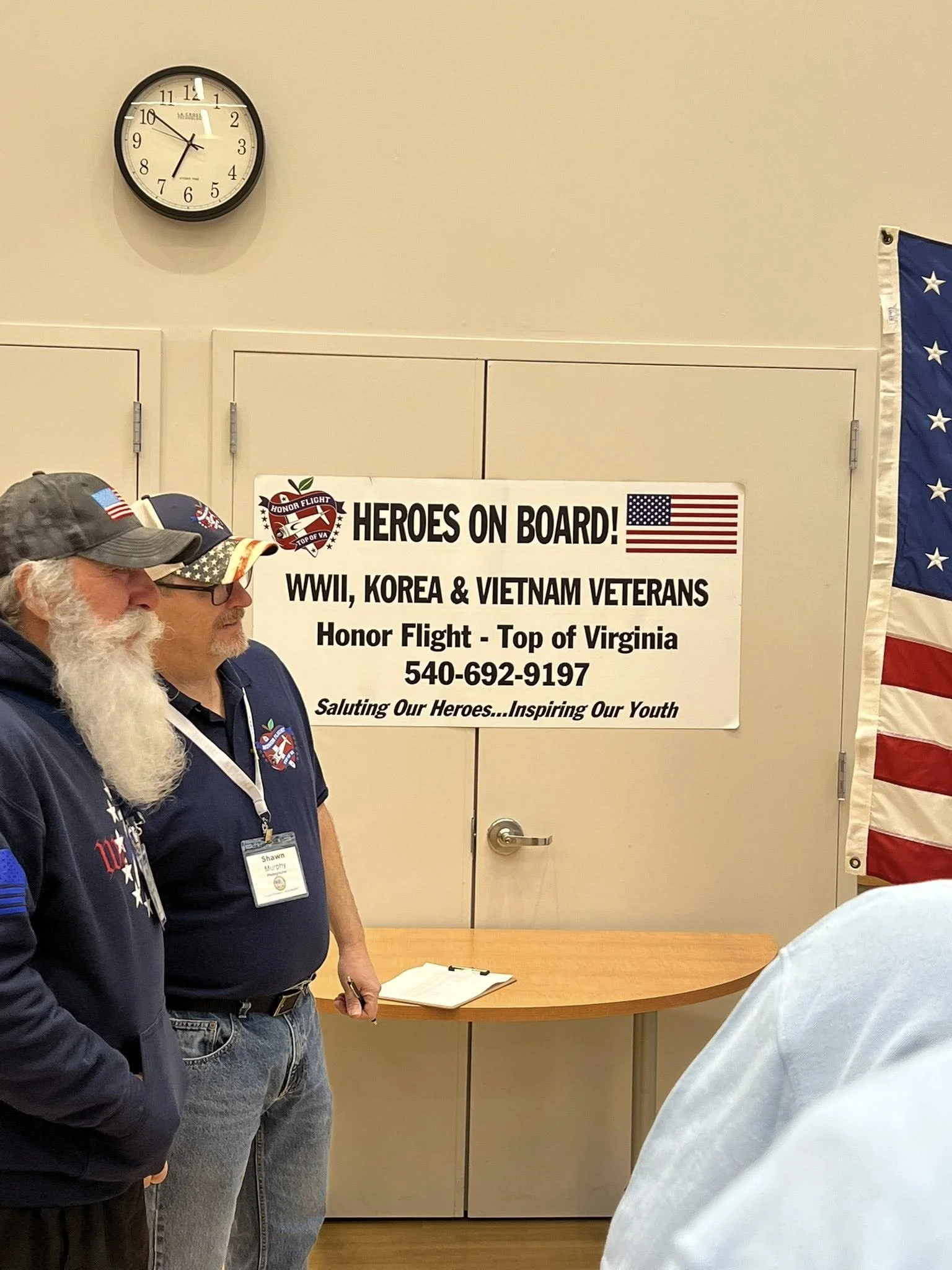 Two men standing in front of a beige wall with a sign that reads "Heroes on Board! WW2, Korea & Vietnam Veterans. Honor Flight - Top of Virginia." One man has a white beard and is wearing a gray cap with an American flag patch, and the other man is w
