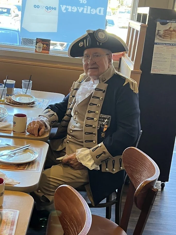 Man dressed as a historical figure in 18th-century military uniform, sitting at a restaurant table.