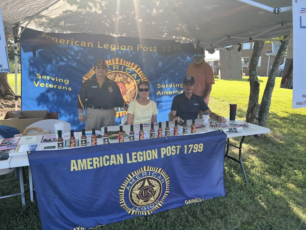 A booth for the American Legion Post 1799 with four people standing or sitting behind it, displaying various hot sauces and informational materials. The booth has a blue banner with the American Legion emblem and text about serving veterans and Ameri