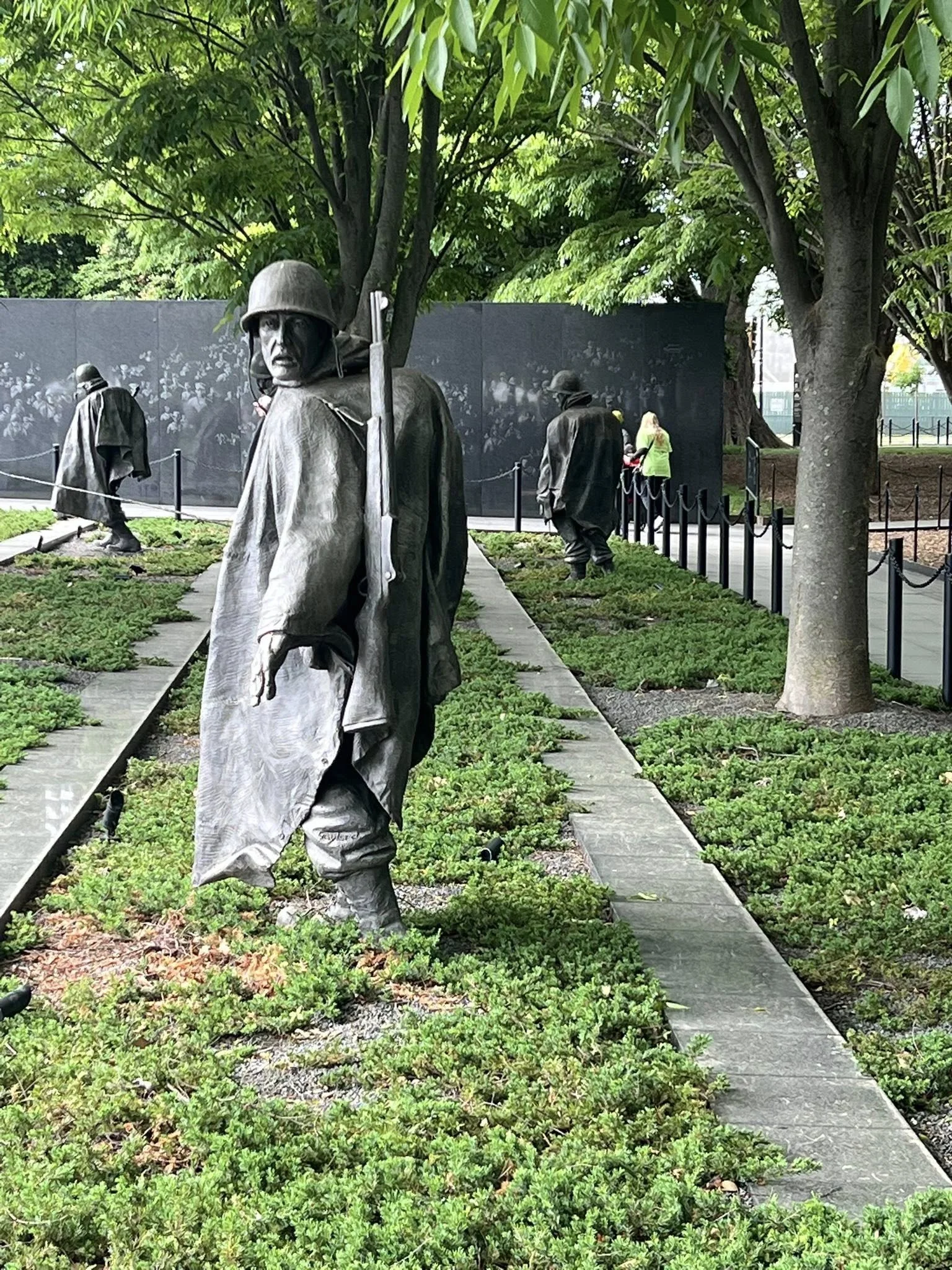 Memorial with bronze statues of soldiers in military uniforms and helmets, set outdoors among trees and shrubs, with a black wall in the background.