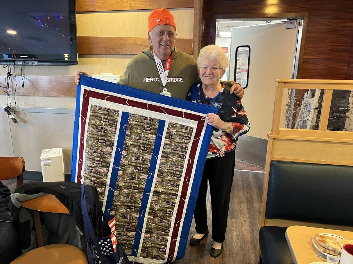 An elderly woman and a man holding a patriotic quilt inside a restaurant. The man wears an orange hat and green hoodie, while the woman wears glasses and a patriotic blouse.