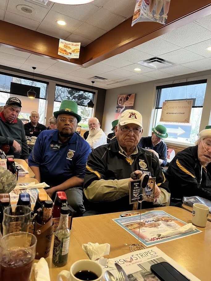 Group of older men sitting at a table in a restaurant, some wearing green and black hats, with a man in the center wearing a beige cap with a red 'M' and a VA (Veterans Affairs) jacket. A couple of men are facing forward, looking at the camera, while