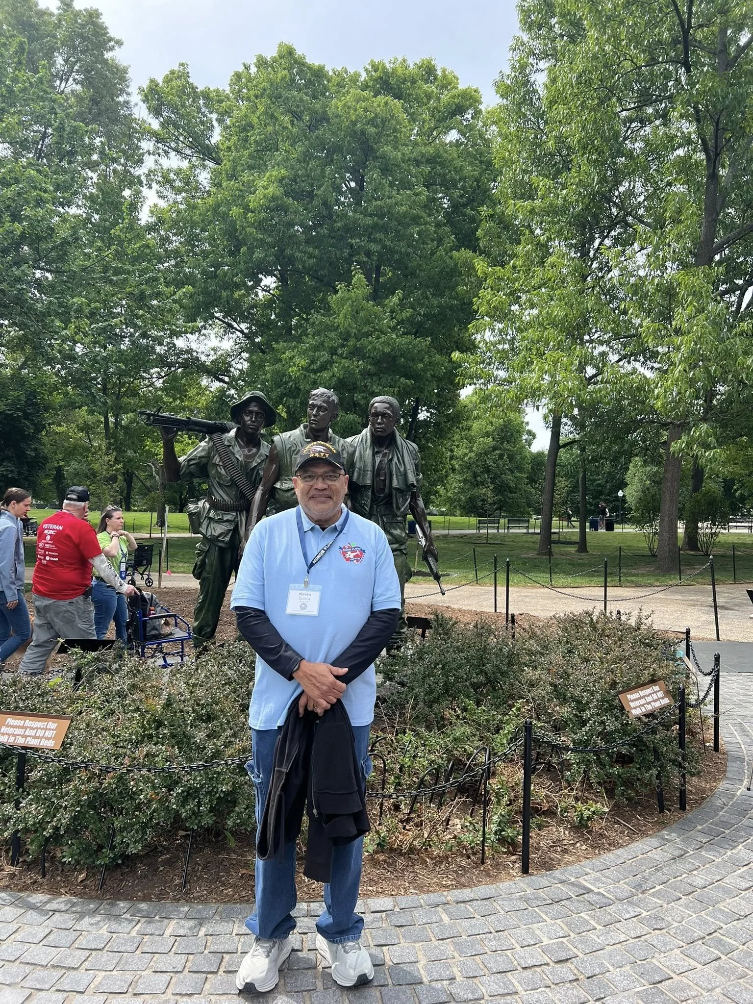 A man standing in front of a war memorial sculpture with three soldiers holding weapons. People are walking in the background in a park with trees and pathways.
