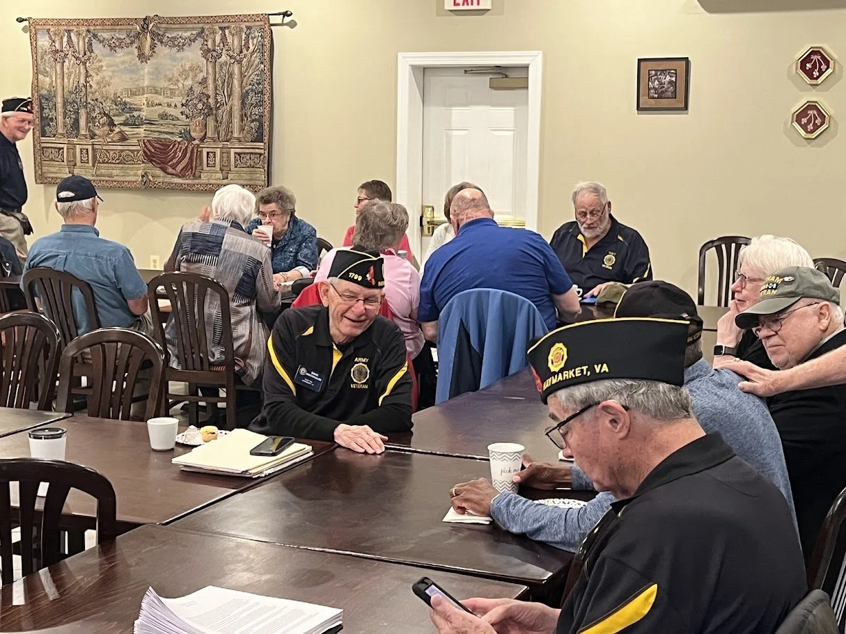 Veterans gathered around a large dining table at a social event, many wearing veteran hats and pins, some engaged with their phones, with a decorative tapestry on the wall behind them.