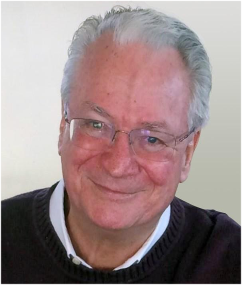 A smiling middle-aged man with gray hair, glasses, and light skin, wearing a dark sweater over a collared shirt.