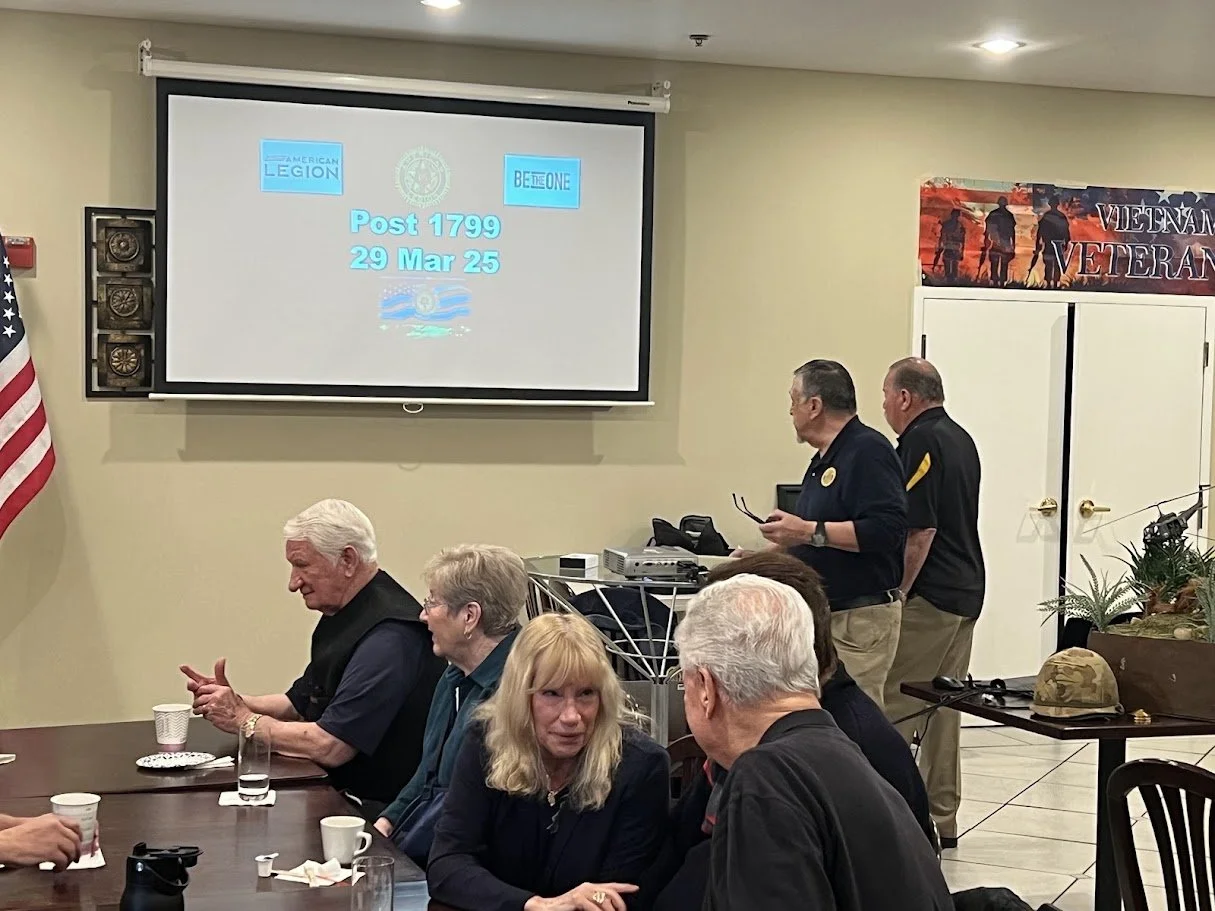 A group of people sitting around a table in a meeting room with a projector screen displaying Post 1799, 29 Mar 25, and logos, with some individuals standing near the wall, and a Vietnam veteran banner in the background.