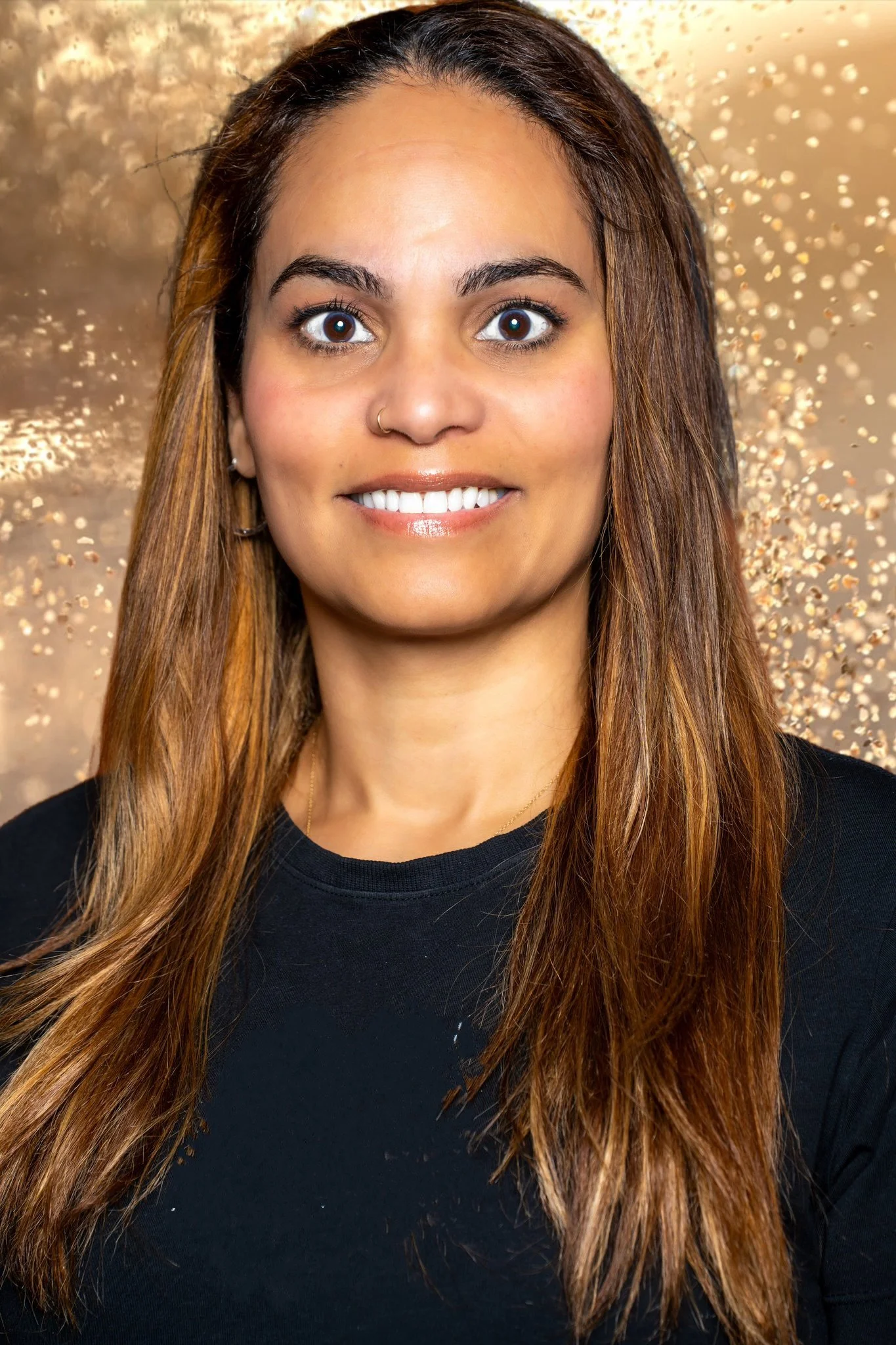 A woman with long brown hair, wearing a black shirt, smiling, with a gold and beige sparkly background.