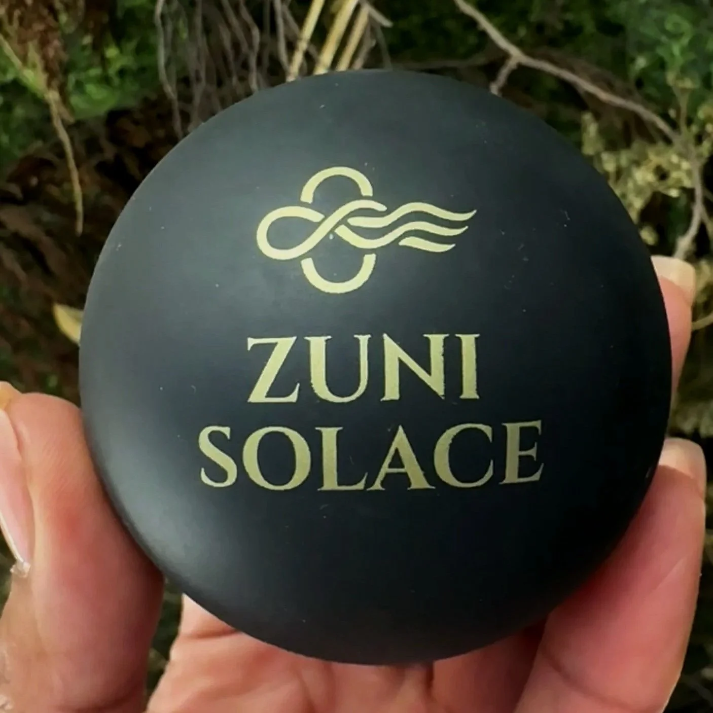 A black round object with gold text that reads 'Zuni Solace' and a gold logo above it, being held by a person's hand outdoors.
