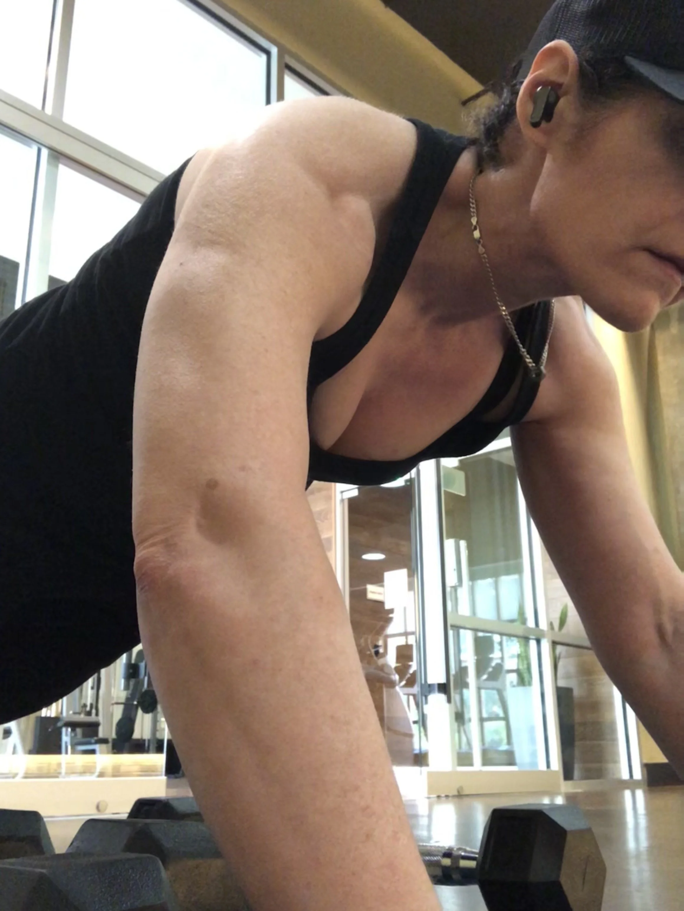 A woman with dark curly hair, wearing a black tank top, exercising on her hands and knees at a gym, with dumbbells and large windows in the background.