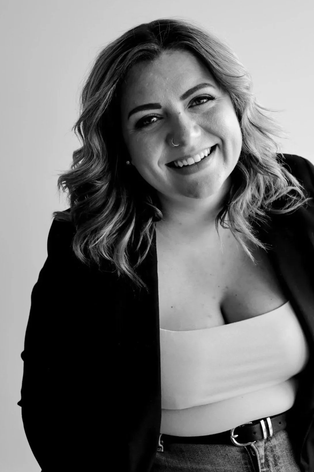 A woman with wavy hair smiling, wearing a light-colored top and a dark blazer, in black and white.