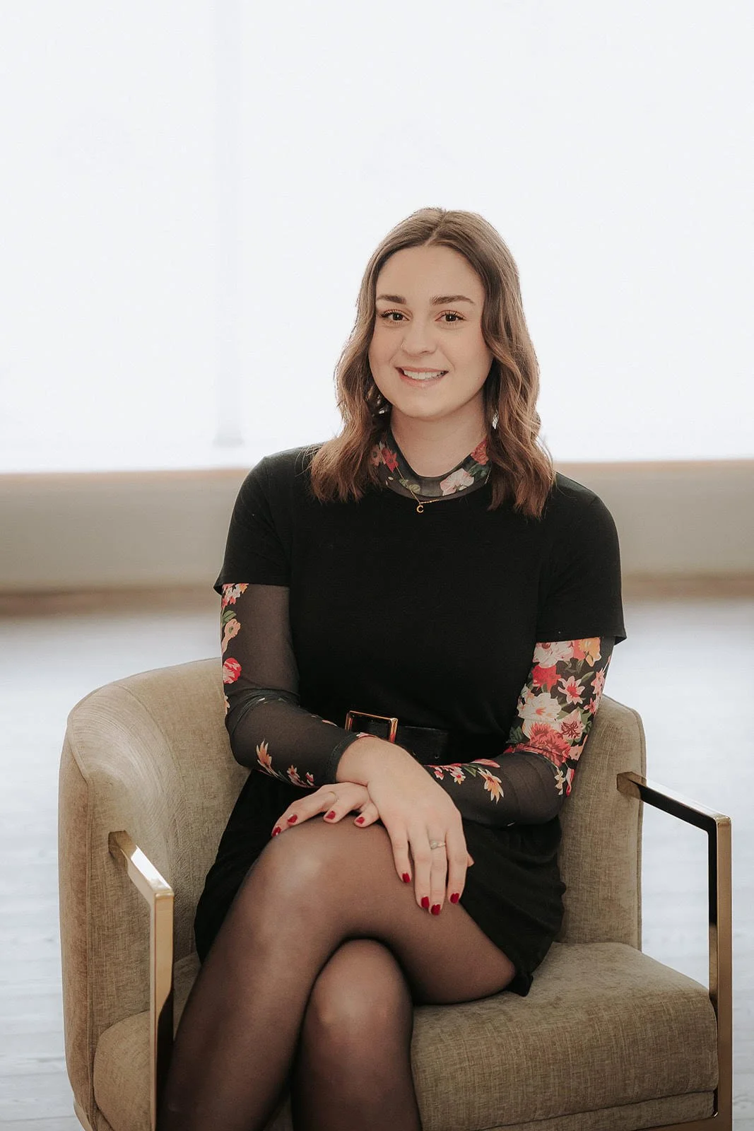 A young woman with shoulder-length brown hair, smiling, sitting on a beige chair with arms crossed over her lap, wearing a black dress with floral patterned sheer sleeves and a floral turtleneck underneath, in front of a bright window.