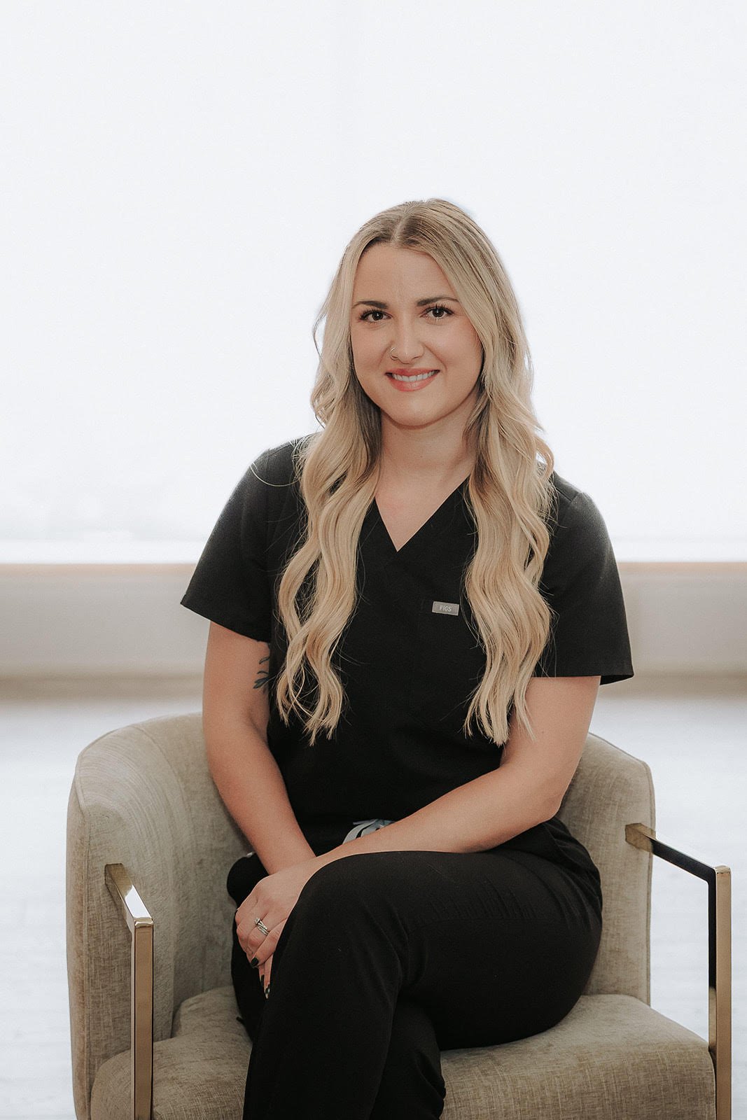 A woman with long blonde wavy hair, wearing a black scrub top with a name badge, sitting on a light-colored armchair indoors.