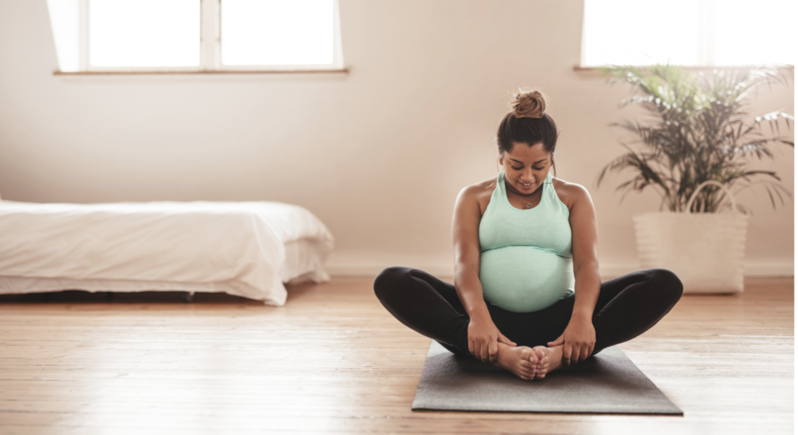 Pregnant woman practicing yoga indoors on a black mat, sitting with legs crossed and holding her feet, with a bed and a potted plant in the background.