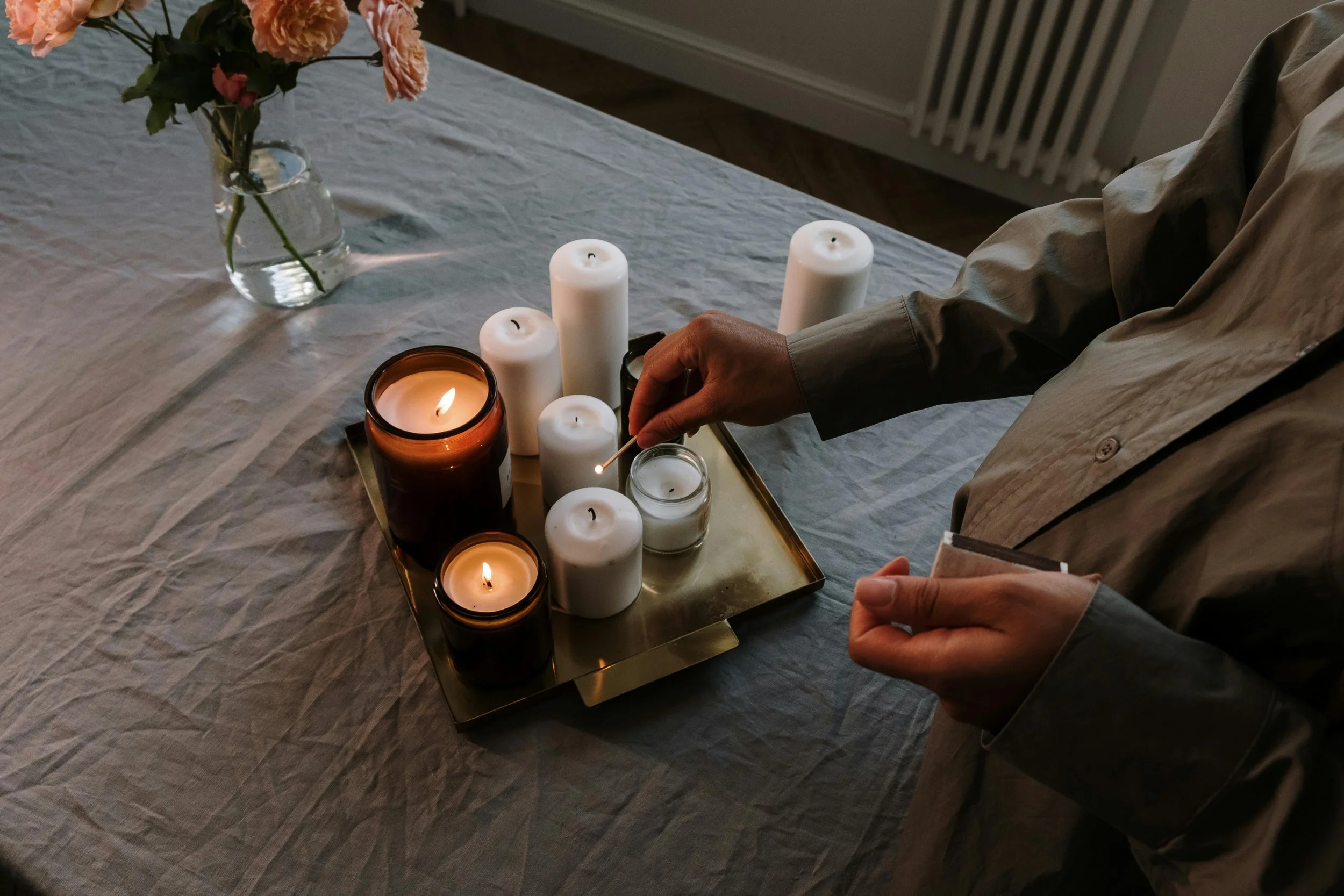 Someone lighting a candle on a tray with various white and brown candles, a matchstick, and a small glass container, on a table with a vase of pink flowers in the background.