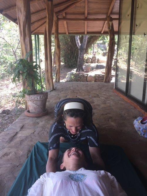 A woman giving a massage to a man lying down on a mat on a porch with wooden beams and a stone floor, surrounded by nature.