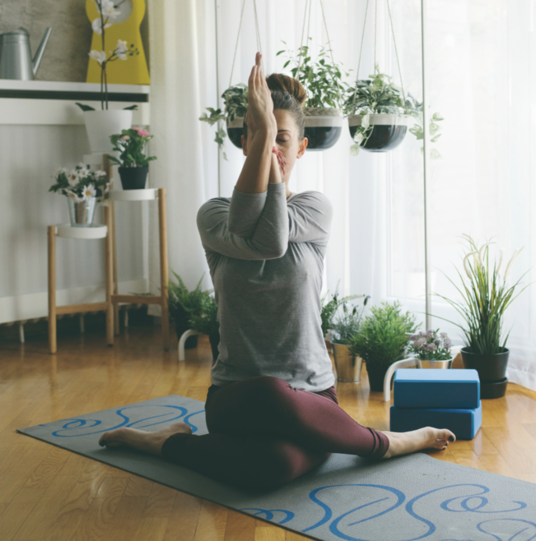 A woman practicing yoga indoors on a mat, sitting in a cross-legged position with her hands pressed together in eagle pose. Practicing yoga while pregnant.