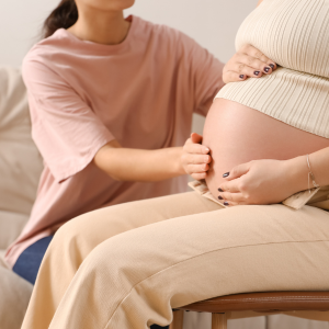 Pregnant woman sitting on a chair with her hand resting on her belly, while another person gently places their hand on her abdomen.