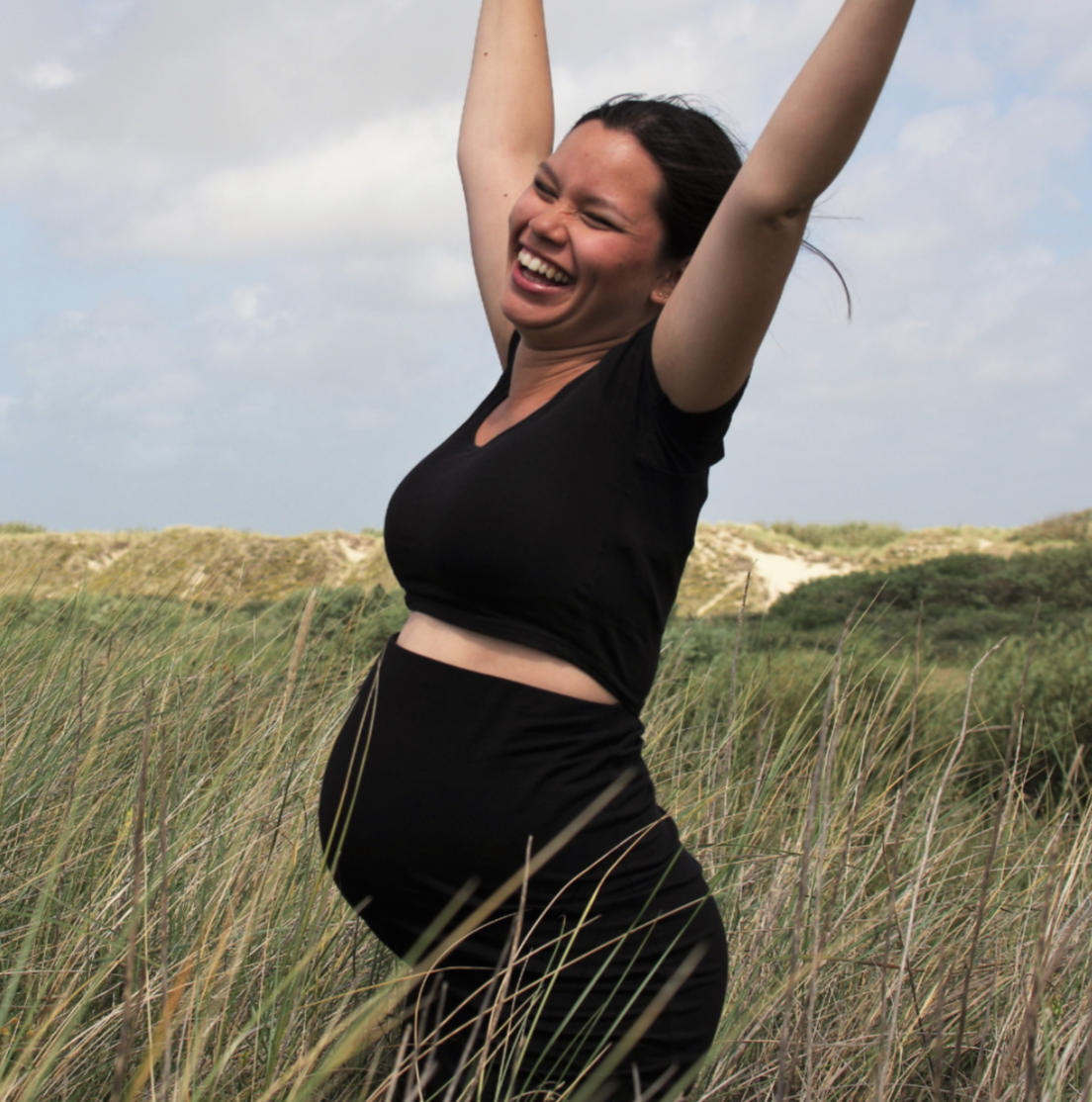 Pregnant woman in black athletic clothes joyfully celebrating outdoors in tall grass with dunes and a cloudy sky.