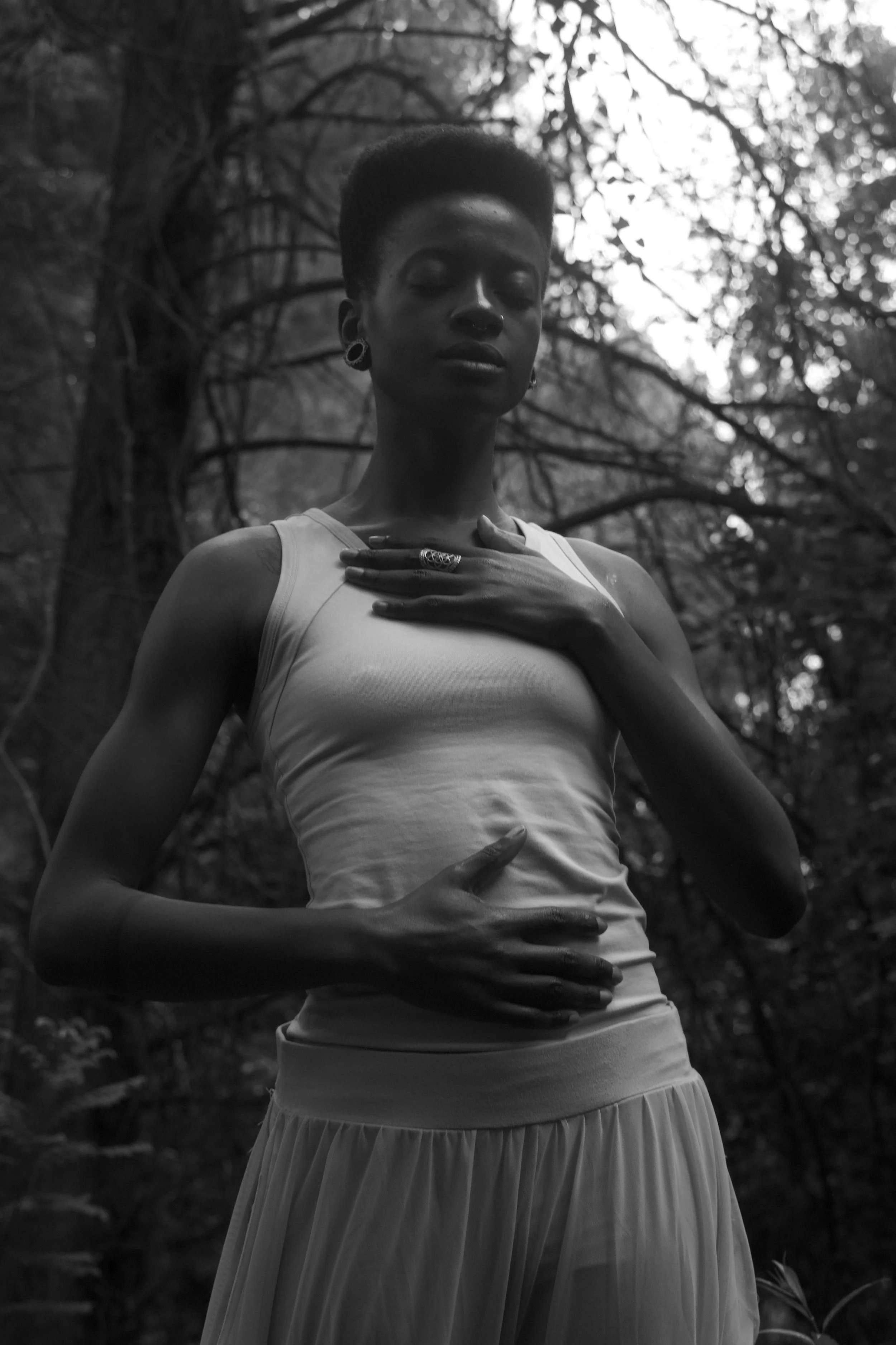 A black and white photo of a woman standing outdoors in a forest, with her eyes closed, wearing a sleeveless top and a pleated skirt, with one hand on her chest and the other on her stomach.