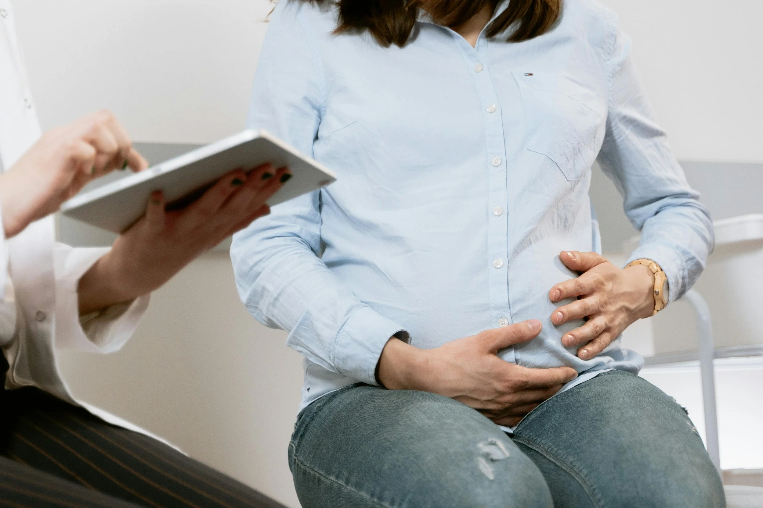 A pregnant woman in a light blue shirt sitting in a medical clinic, with hands on her belly, during a consultation with a healthcare professional.