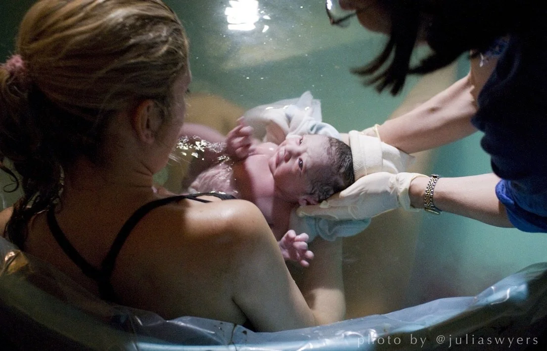 A woman giving birth in a birthing pool while a medical professional assists with the delivery, holding the newborn baby in the water.