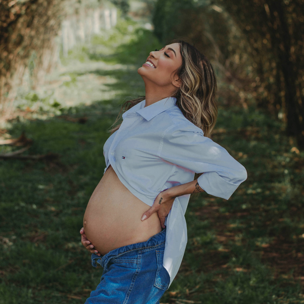 A pregnant woman outdoors, smiling joyfully with her hand on her belly, wearing a light blue shirt and jeans in a green park setting.
