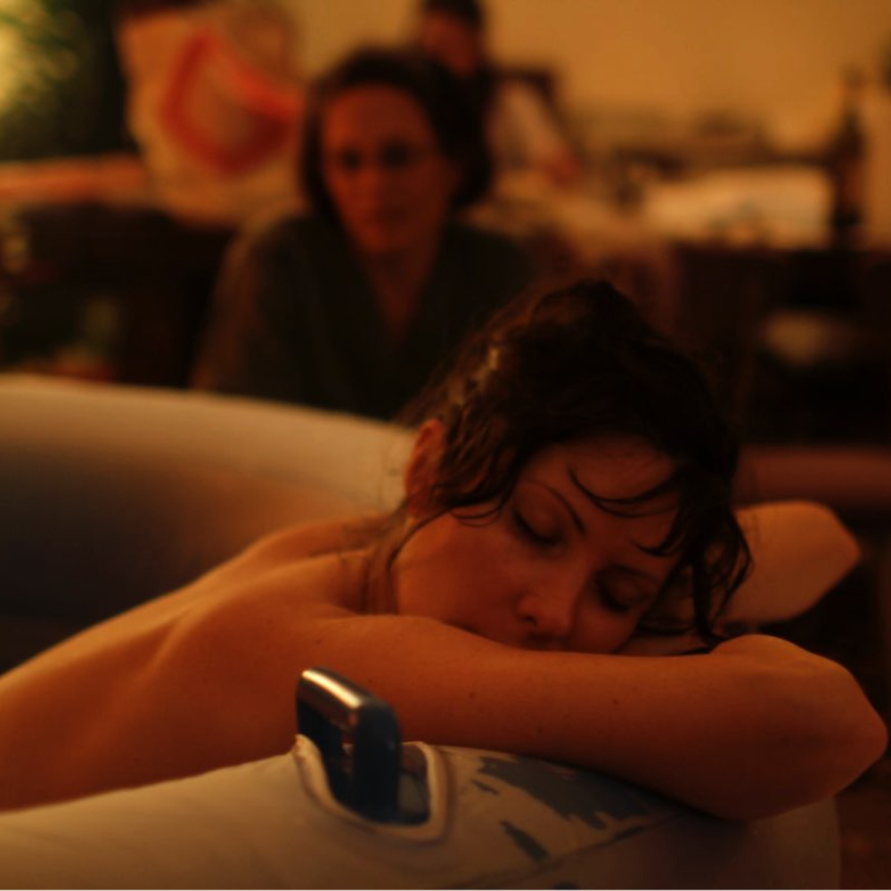 A woman with dark hair sleeping on her side with her arms crossed on a bed in a dimly lit room. A phone is placed on the bed near her arm.