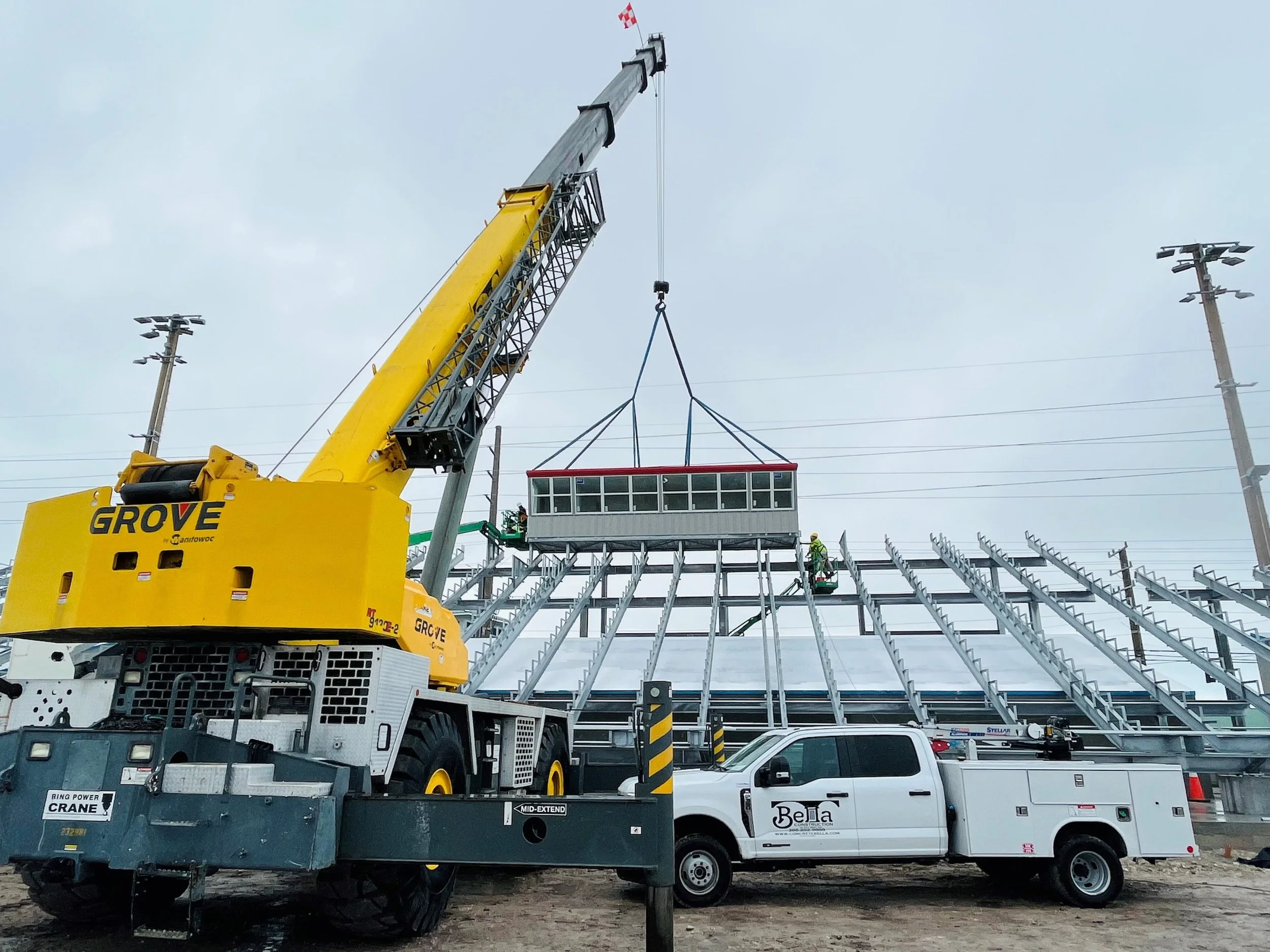 Construction workers installing a glass enclosure on a steel structure with a large yellow crane and a white utility truck on site.