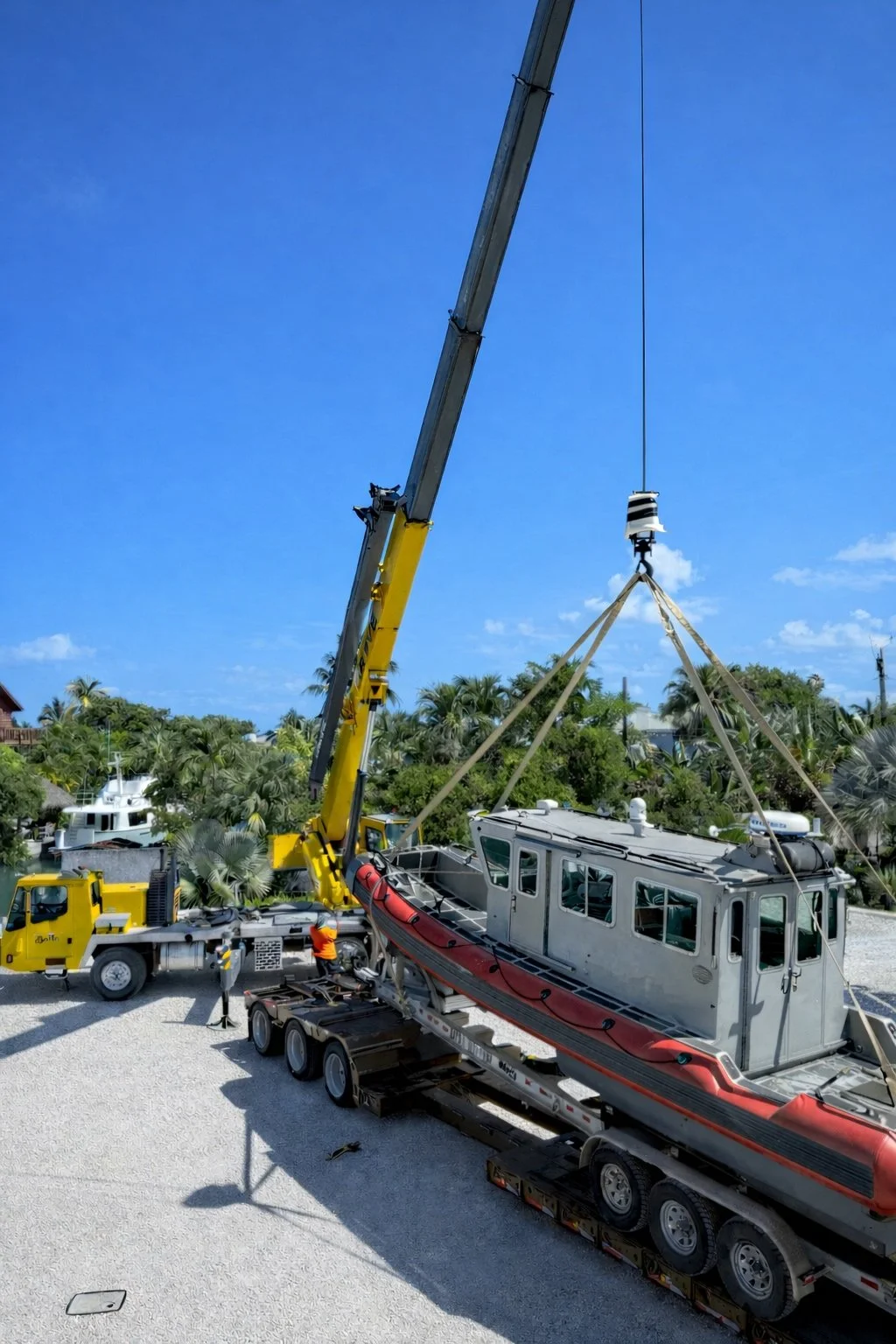 A crane lowering a small boat with a cabin onto a trailer in a yard with palm trees and blue sky.