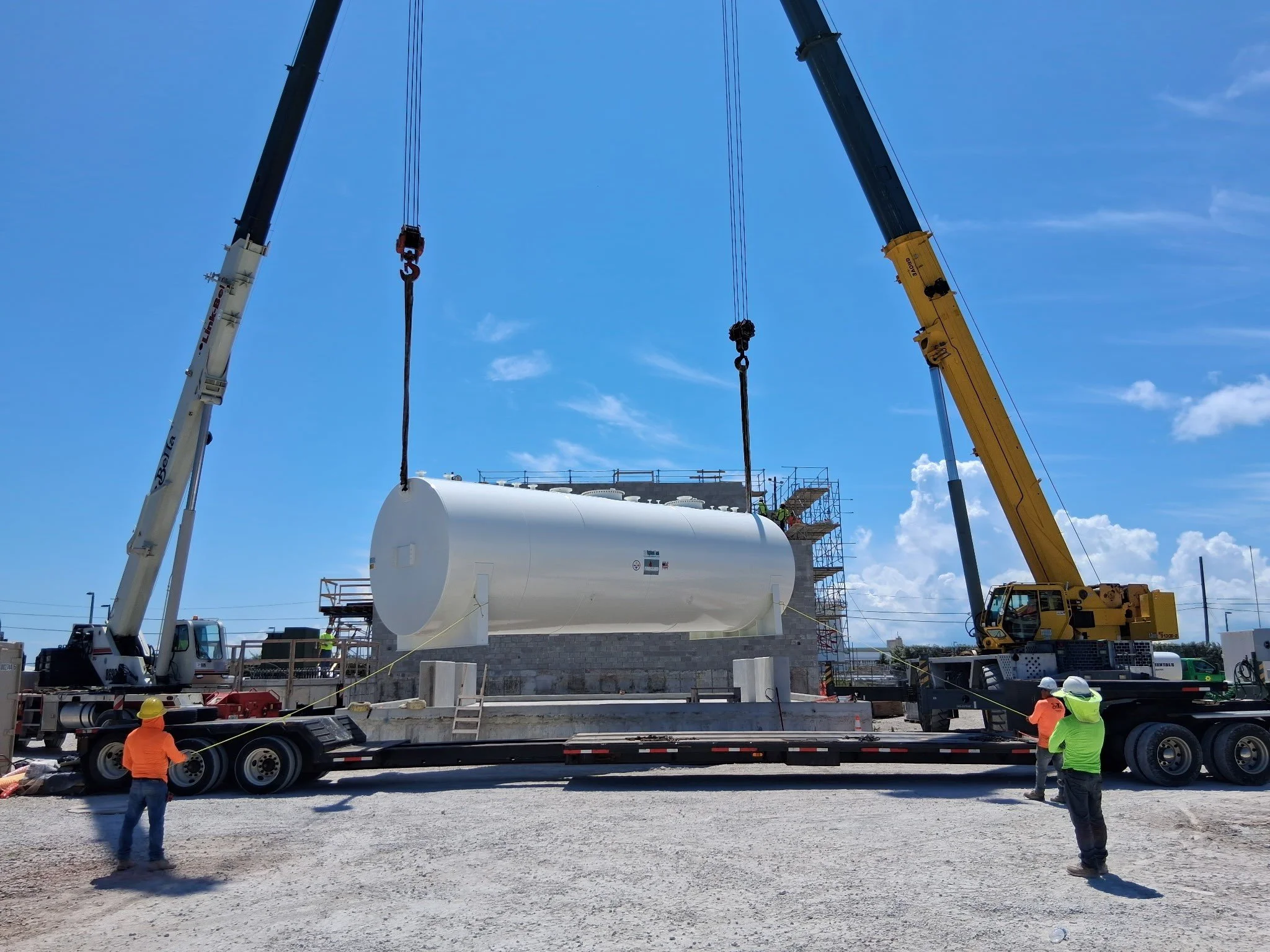Two cranes lifting a large white cylindrical tank into place at a construction site with workers overseeing the operation under a blue sky.