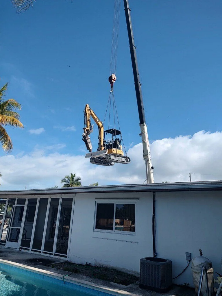 A construction crane lifting a small excavator above a house with a pool in the foreground and palm trees in the background.
