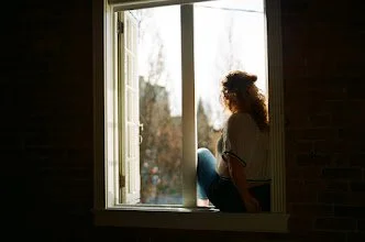 A woman sitting by a window looking outside, with sunlight illuminating her silhouette.