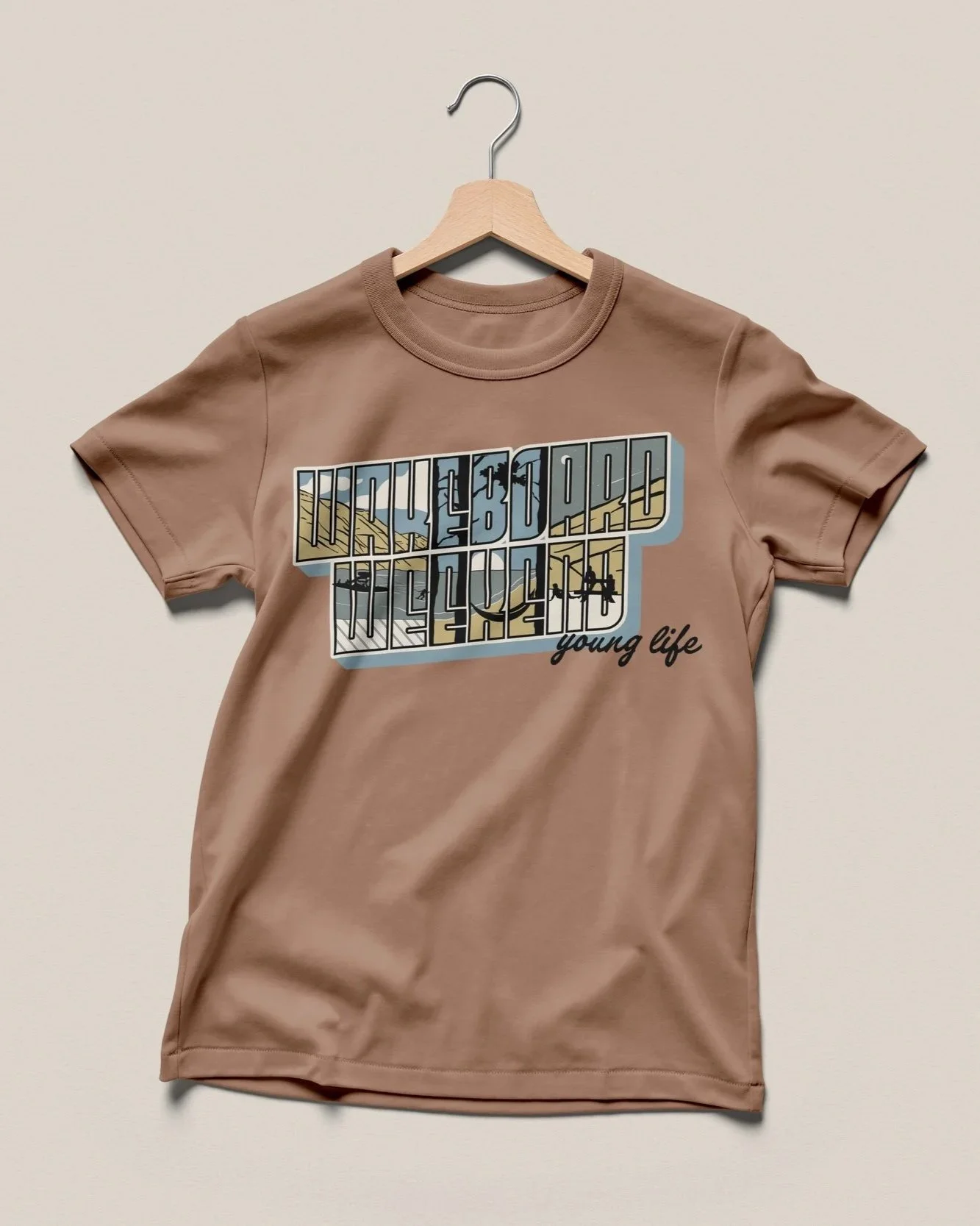 A beige T-shirt hanging on a wooden hanger on a beige wall, with a graphic design that includes mountains and text.