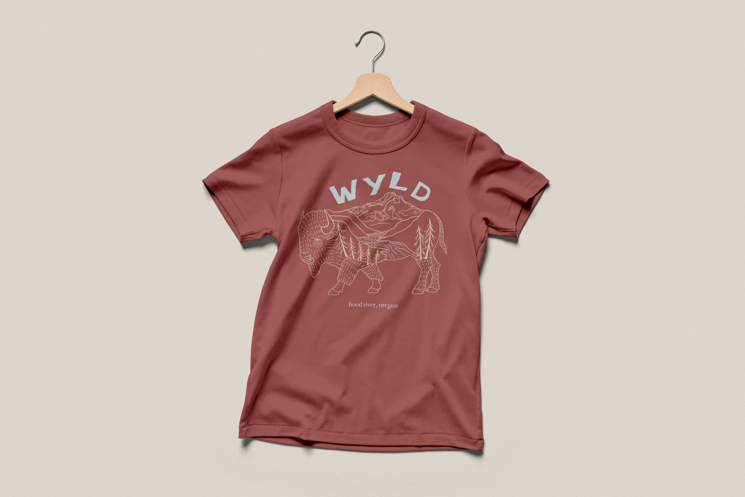 A pink T-shirt hanging on a wooden hanger against a neutral background. The T-shirt features an outline graphic of a bison overlaid with a mountain and forest scene, with text that says "WYLD" and "hood river, oregon" underneath.