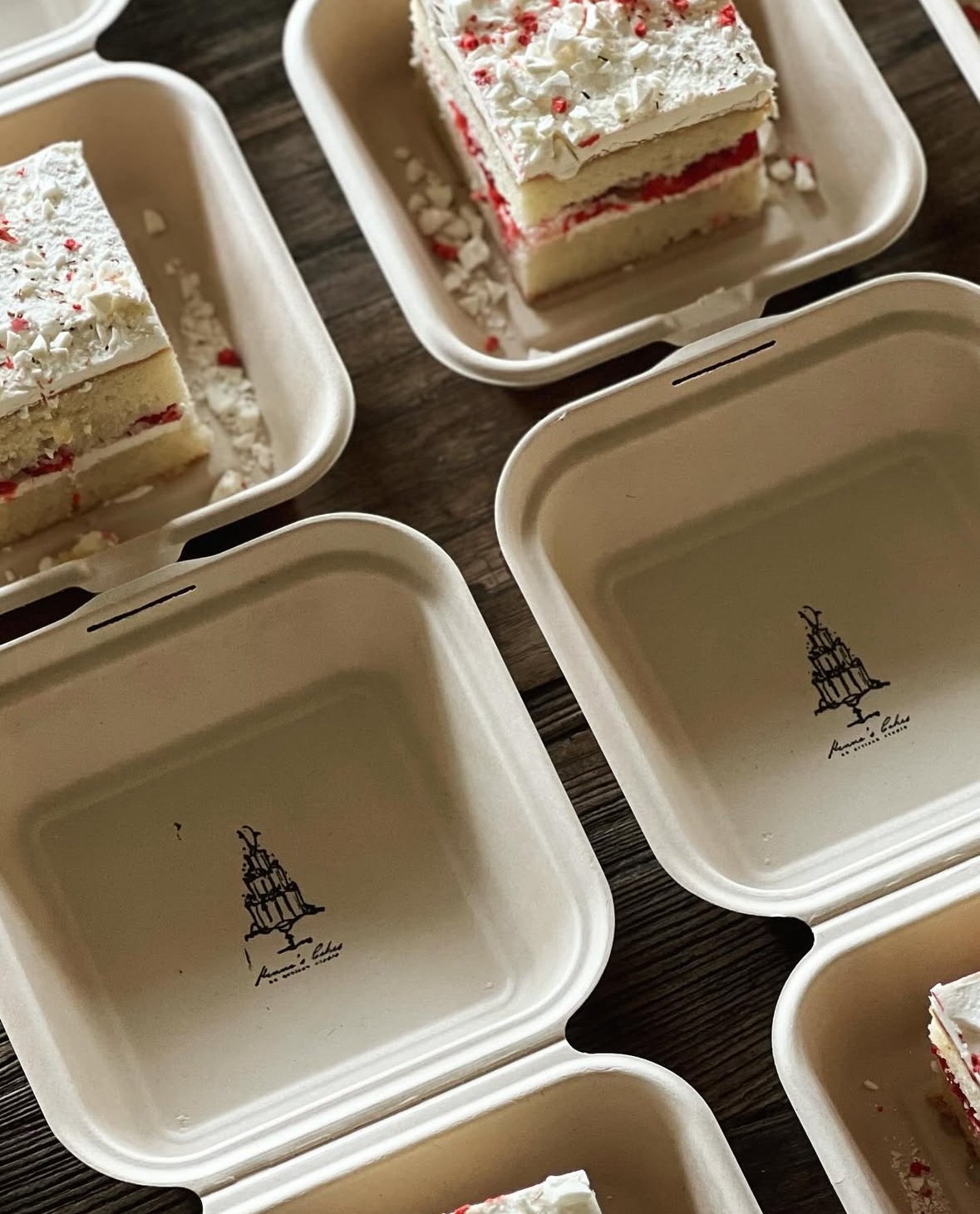 Empty white takeout containers with a logo of a layered cake. Some contain slices of layered cake with white frosting, red filling, and sprinkles on top, placed on a wooden surface.